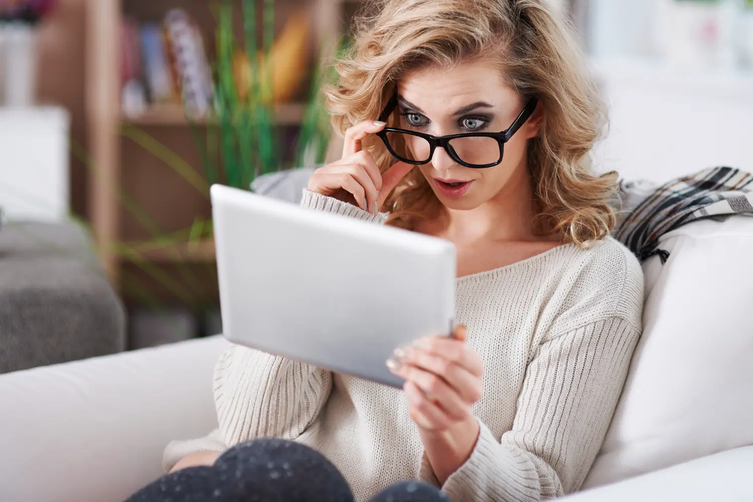 Woman reading a tablet with glasses in a cozy home setting.