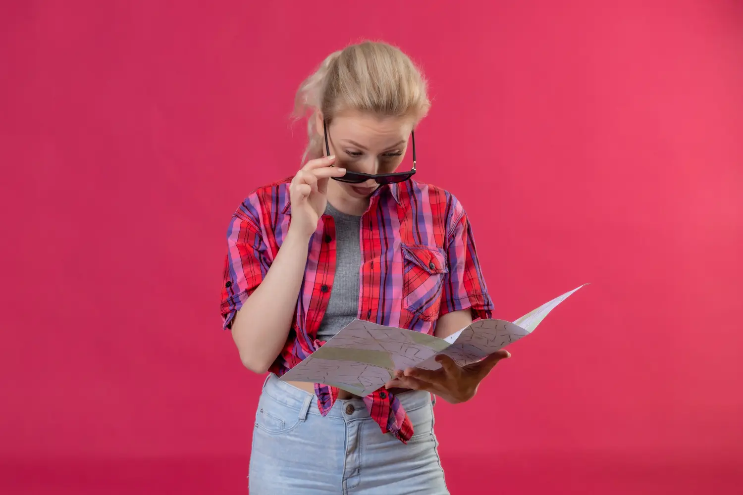 Woman reading a map with sunglasses against a pink background.