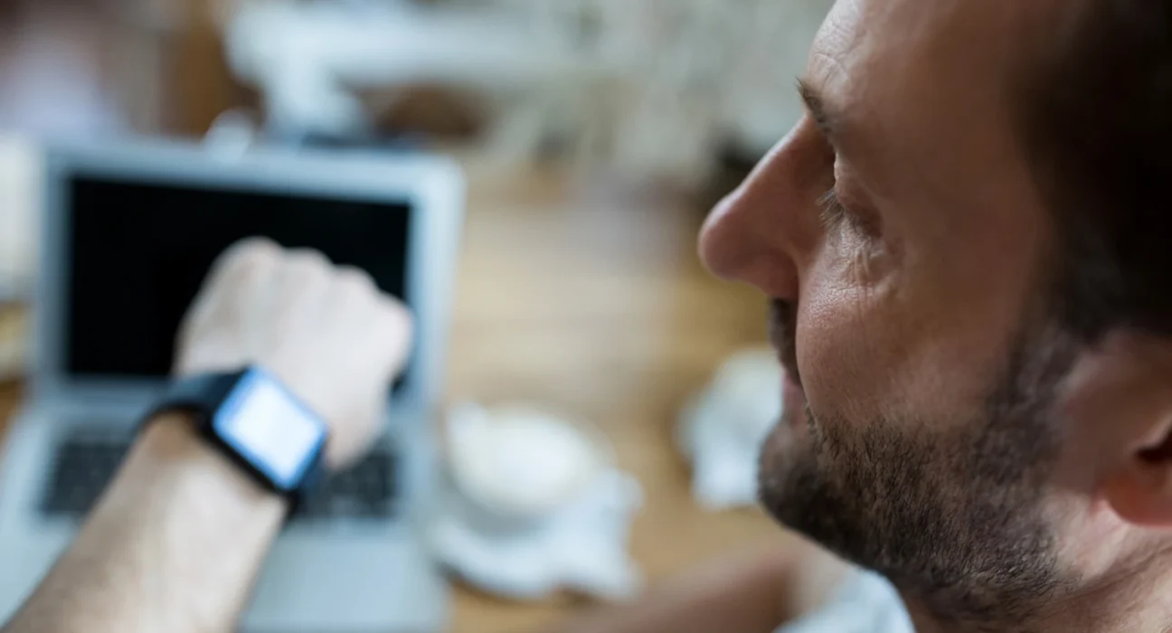Man looking at his smartwatch while working on a laptop, emphasizing the importance of time manageme.