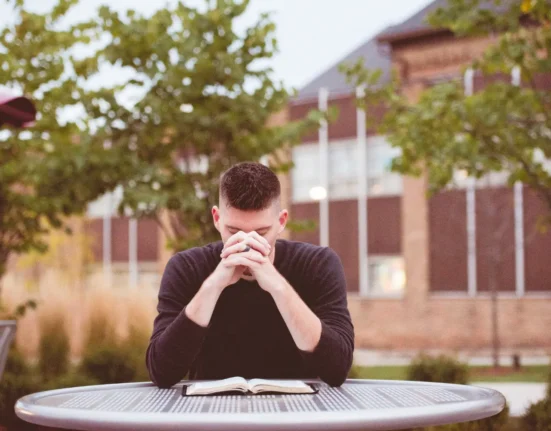 Student studying outdoors with hands clasped over face, sitting at a table with an open book.