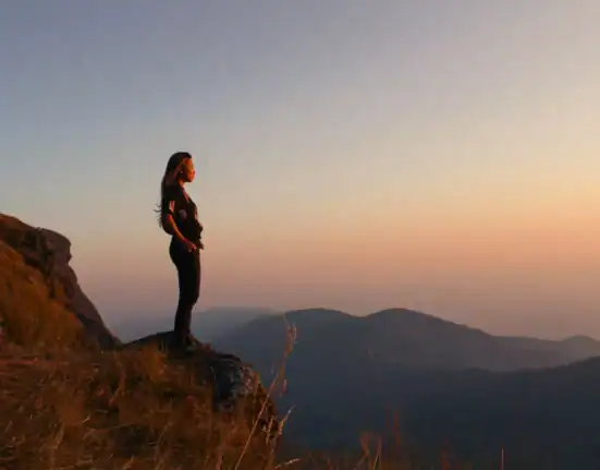 A woman standing on a mountain edge during sunset, contemplating the scenic landscape.
