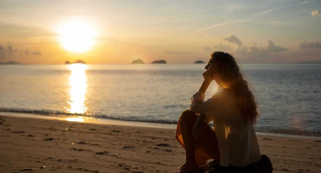 Sunset reflection on the ocean with a woman sitting on the beach, embracing self-love and personal g.