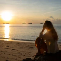 Sunset reflection on the ocean with a woman sitting on the beach, embracing self-love and personal g.