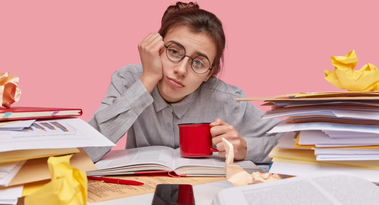 Frustrated woman overwhelmed with paperwork and a coffee mug at her cluttered desk.
